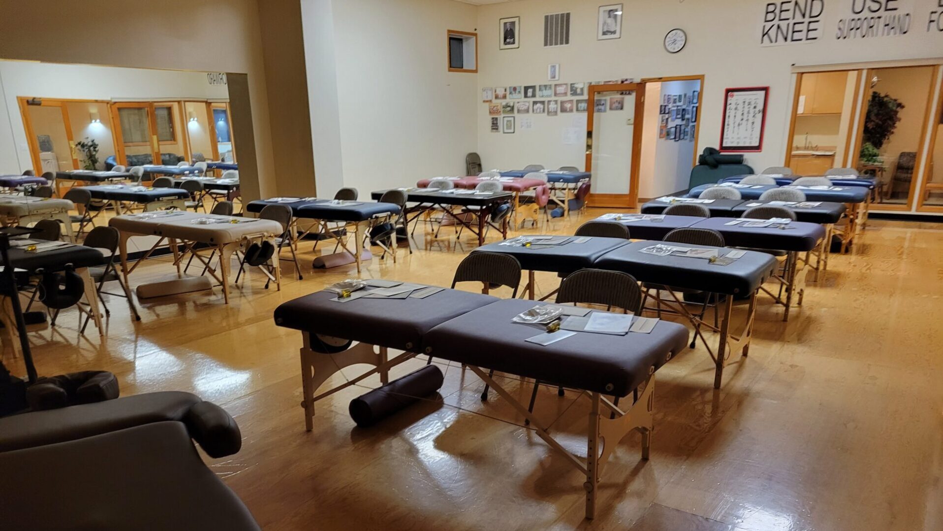 A sparsely populated community hall set up with tables and chairs for an event, featuring a massage table with papers on it in the foreground.